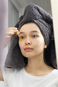 An Asian woman applies skincare serum with a dropper in front of a mirror.