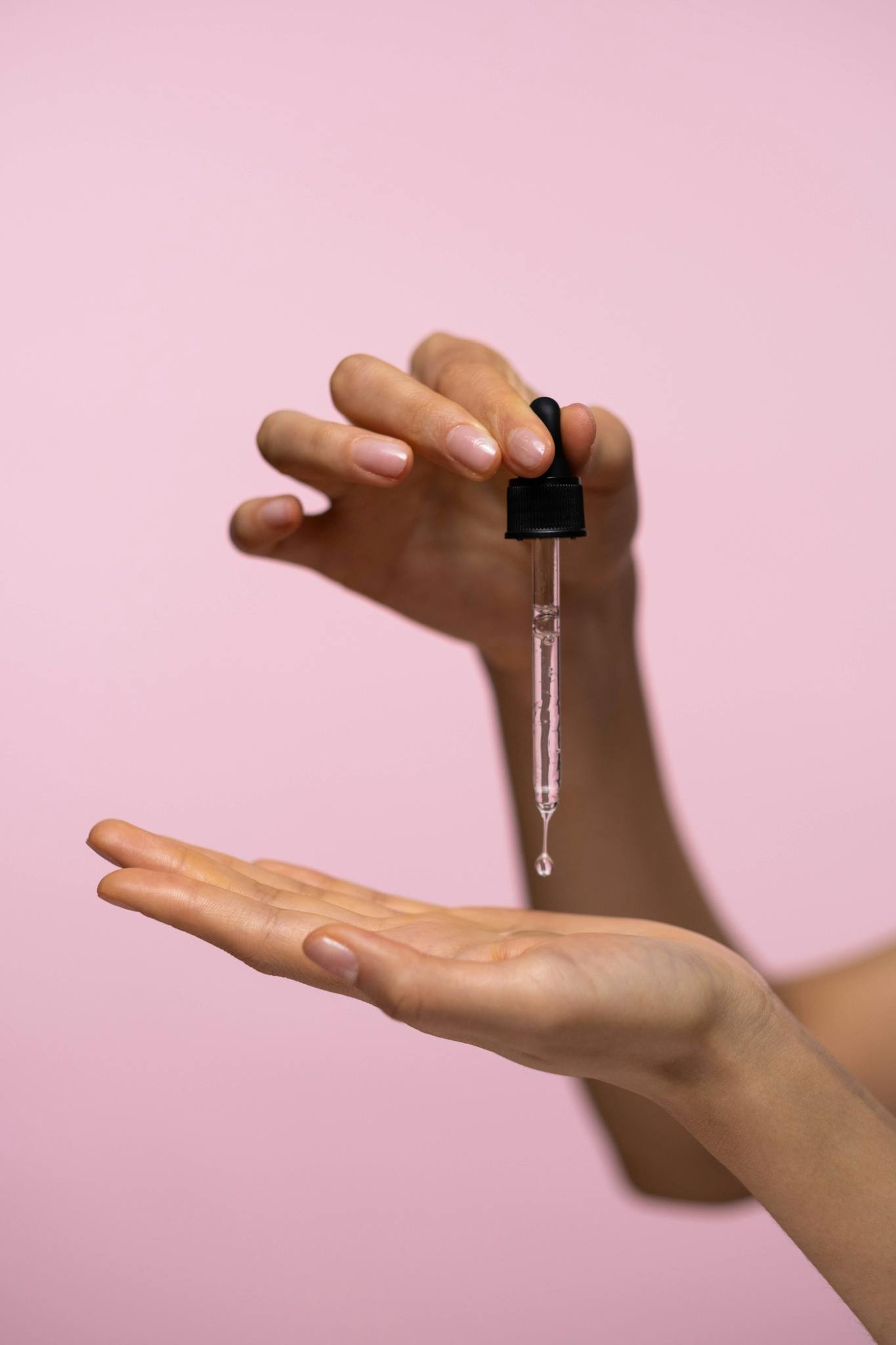 Close-up of hands with dropper applying face serum against a pink background for skincare routine.